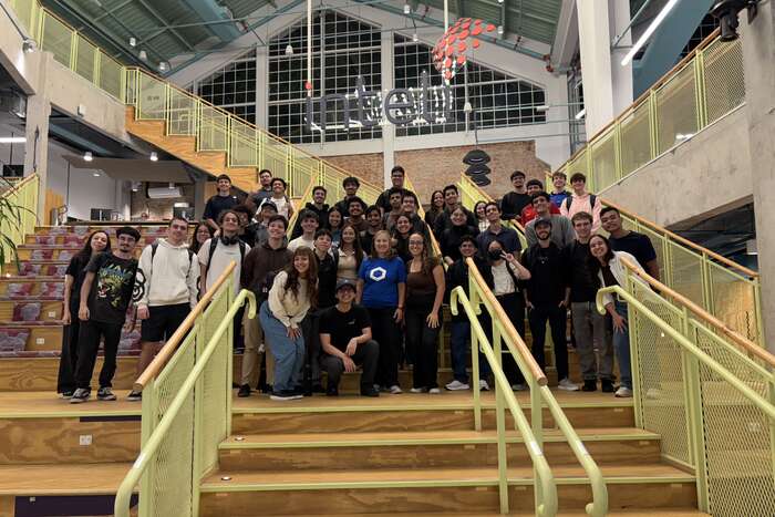 About forty-five bootcamp participants posing on the central staircase at Inteli, with the school signage on the wall behind them.