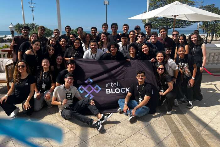 About forty Inteli Blockchain members gathered outdoors by the sea, in club t-shirts and badges, holding the club flag.
