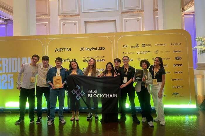 Ten club members on the Meridian 2025 stage holding the Inteli Blockchain flag, two of them with trophies in hand, in front of the event sponsor wall.