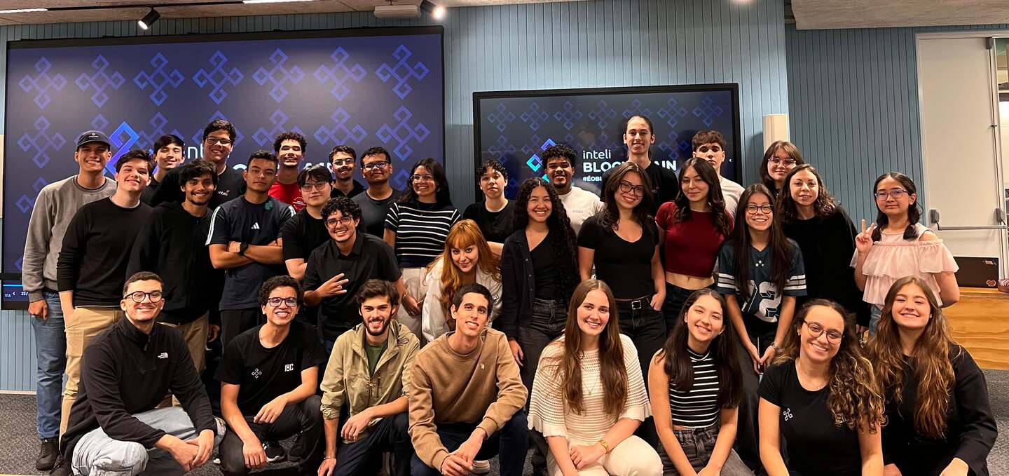 About thirty-five Inteli Blockchain members posing together in two rows in an Inteli auditorium, with the screens behind them showing the club logo.