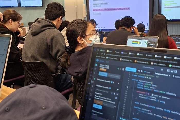 An Inteli classroom during the workshop: students in rows with open laptops, projection screens in the background and, in the foreground, a laptop with contract code open in the editor.